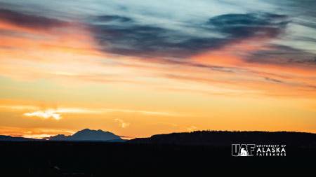 Dark hills in the foreground with an orange to blue gradient sunset illuminates the silhouette of Denali in the background, and the UAF logo in the lower right corner.