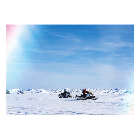 Photo of person on a snowy landscape driving a snowmobile that is pulling equipment behind it. A color sundog overlay highlights the corners on the photo.
