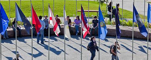 Students walking through campus on beautiful, sunny day. Flagpoles stand in the forefront of the photo.