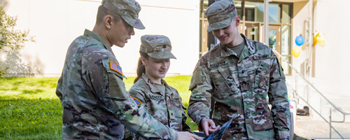 The classroom setting features a group of individuals, predominantly in military attire, engaged in a lesson. A teacher stands at the front, utilizing a whiteboard filled with mathematical equations.