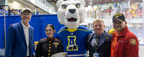 A service member dressed in military fatigues walks confidently along a snowy path on campus. She holds a laptop and a textbook in her arms while a backdrop of trees and a church steeple can be seen under a clear blue sky.